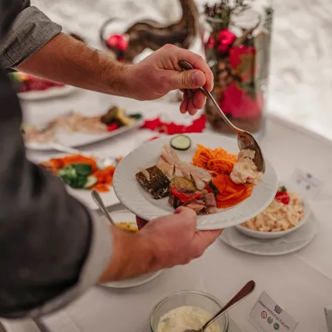 Person, die sich an einem Buffet den Teller füllt und mit einem Löffel Speisen auf einen weißen Teller gibt, auf dem bereits eingelegtes Gemüse, geraspelte Karotten, Gurkenscheiben, marinierter Fisch und Aufschnitt liegen. Der Buffettisch ist mit Schüsseln mit Salaten, Saucen und Brot gedeckt und mit roten Weihnachtsornamenten und Kerzen dekoriert, um eine festliche Atmosphäre zu schaffen.