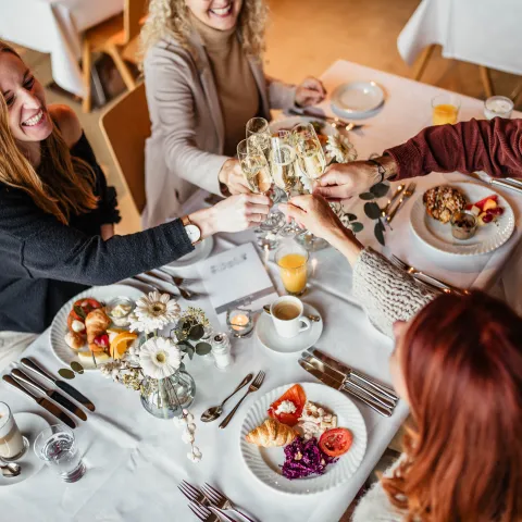 Eine Gruppe von Freunden genießt einen Brunch in einem Restaurant und erhebt ihre Champagnergläser zu einem Toast über einer weißen Tischdecke. Es werden Croissants, frisches Obst, Tomaten, Rotkohlsalat, Müsli mit Joghurt und Räucherlachs serviert, dazu Kaffee, Orangensaft und Latte Macchiato. Der Tisch ist mit einer Vase mit weißen Blumen dekoriert, was eine festliche und elegante Atmosphäre schafft