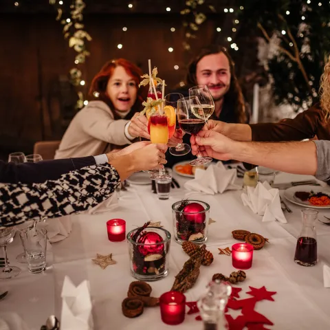 Eine Gruppe lachender Menschen sitzt an einem weihnachtlich dekorierten Tisch und stößt feierlich mit Getränken an
