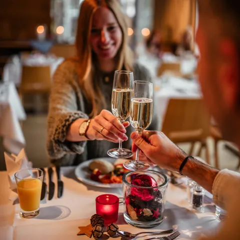 Zwei Personen stoßen mit Sektgläsern an, während sie an einem festlich gedeckten Tisch sitzen. Auf dem Tisch sind ein Teller mit Essen, ein Glas Orangensaft und eine dekorative Glasvase mit roten und schwarzen Elementen zu sehen.