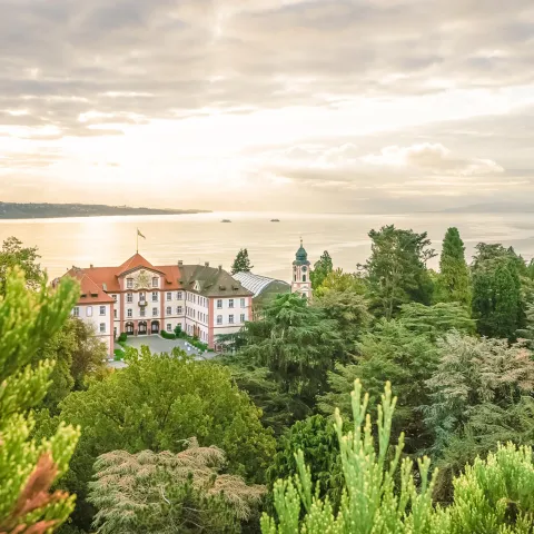 Blick über eine grüne Parklandschaft auf ein historisches Gebäude am Ufer eines Sees, im Hintergrund ruhiges Wasser und bewölkter Himmel im Abendlicht.