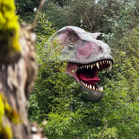 Kopf eines Dinosauriermodells mit geöffnetem Maul und scharfen Zähnen, der zwischen dichtem grünen Gebüsch hervorschaut