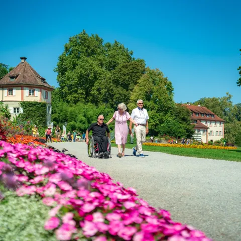 Drei ältere Menschen spazieren gemeinsam durch einen Park, eine Person im Rollstuhl, umgeben von bunten Blumenbeeten und historischen Gebäuden