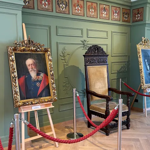 Ausstellungsraum zur Ausstellung „200 Jahre Großherzog Friedrich“ mit einem historischen Holzstuhl, zwei gerahmten Porträts auf Staffeleien und rot abgesperrtem Bereich vor grüner Wandvertäfelung. An der oberen Wand sind mehrere Wappen und Namensschilder angebracht.