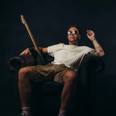 Person mit Sonnenbrille sitzt lässig in einem Ledersessel, neben sich eine E-Gitarre, vor dunklem Hintergrund
